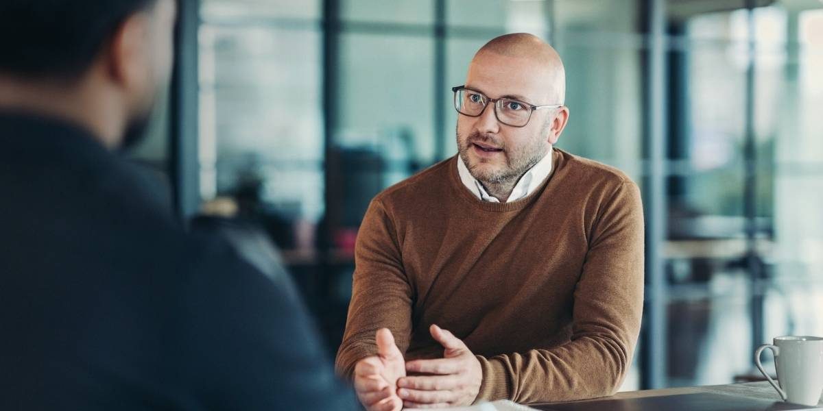 Glatzköpfiger Berater mit Brille und Bart im braunen Pullover spricht gestikulierend mit einer Person in einem modernen Büro. Eine weiße Tasse steht auf dem Tisch.