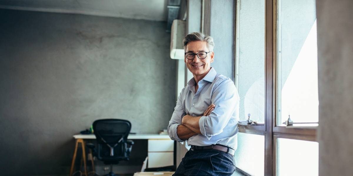 Glatzköpfiger Berater mit Brille und Bart im braunen Pullover spricht gestikulierend mit einer Person in einem modernen Büro. Eine weiße Tasse steht auf dem Tisch.