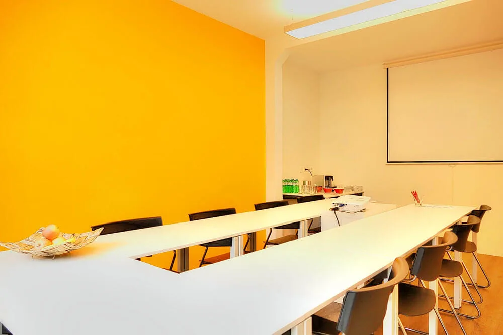Modernes Besprechungszimmer mit U-förmigem weißen Tisch, schwarzen Stühlen, gelber Wand und ausfahrbarer Leinwand.