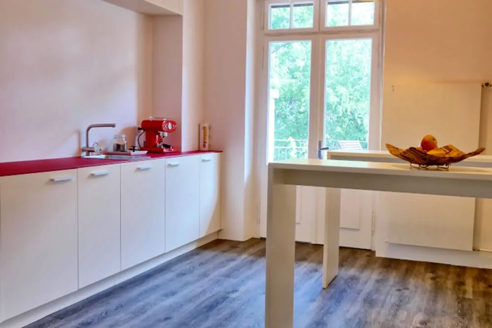 Moderne Küchenzeile mit roter Espressomaschine, Spüle, weißen Schränken und roter Arbeitsplatte. Großer Fensterblick, heller Essbereich.