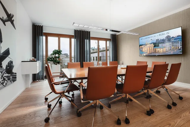 Moderner Konferenzraum mit braunem Holztisch, Ledersesseln, großen Fenstern, TV-Bildschirm mit Berlin-Motiv und Kunst an der Wand.