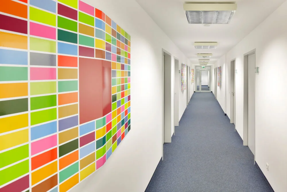 Langer, heller Flur mit blauem Teppichboden und weißen Wänden, geschmückt mit einer großen, bunten Mosaik-Wanddekoration.