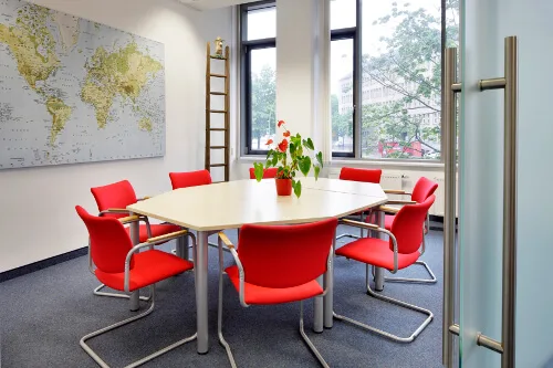 Heller Besprechungsraum mit achteckigem Tisch, acht roten Stühlen, großer Weltkarte und Fensterblick auf Stadt.