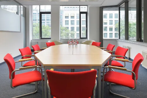 Ein heller Konferenzraum mit einem achteckigen Holztisch, umgeben von roten Stühlen und großen Fenstern mit Stadtblick.