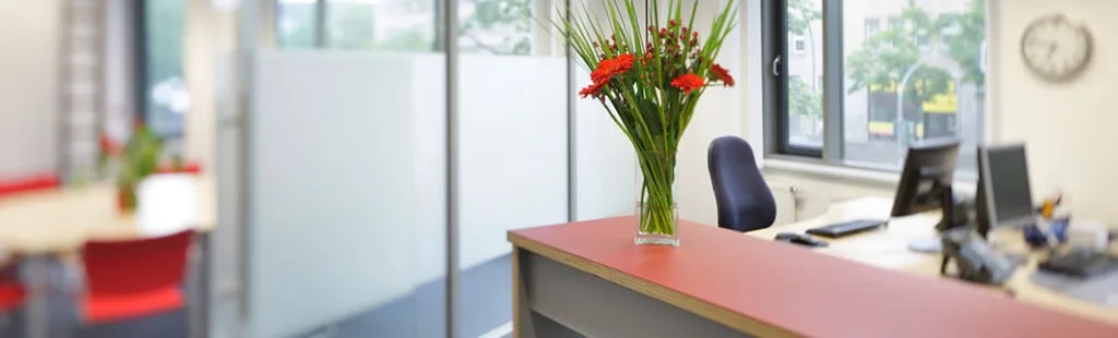 Moderner Empfangsbereich eines Büros mit rotem Tresen, frischen roten Blumen in einer Vase und dahinterliegenden Arbeitsplätzen.