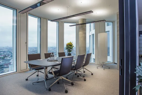 Moderner Konferenzraum mit großem Tisch, dunklen Stühlen, Blick auf Stadtpanorama durch große Fenster, Teppichboden und modernen Leuchten.