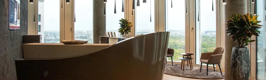 Moderne Rezeption mit geschwungener Theke vor Panoramafenstern, die einen weiten Blick auf Stadt und Park bieten, ergänzt durch stilvolle Pflanzen und Sitzgelegenheiten.