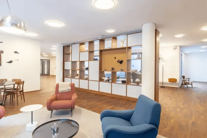 Geräumige, modern eingerichtete Lounge oder Gemeinschaftsbereich mit roten und blauen Sesseln, Couchtischen, einem Holzregal und Parkettboden.