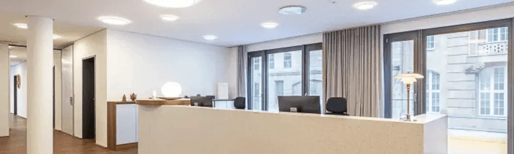 Heller, moderner Empfangsbereich mit langer Rezeption, Computern, runder Designlampe und großen Fenstern zu einem historischen Gebäude.