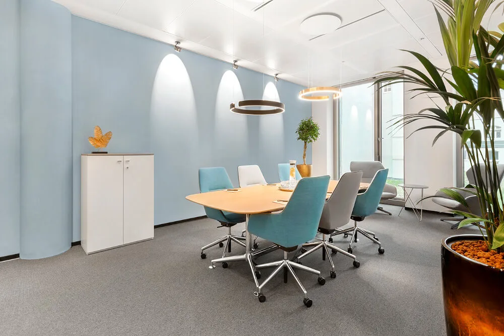 Moderner Besprechungsraum mit hellblauen Wänden, großem Holztisch, blauen Stühlen, grauem Teppichboden, Pflanzen und Design-Leuchten.