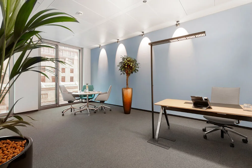 Moderner Büroraum mit blau getönter Wand, großer Fensterfront, Meetingbereich mit Rundtisch, Schreibtisch und Stehleuchte.