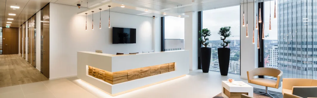 Moderner Empfangsbereich in einem Büro mit weißem Tresen, TV, Pendelleuchten, großer Fensterfront mit Stadtblick und Sitzgelegenheiten.