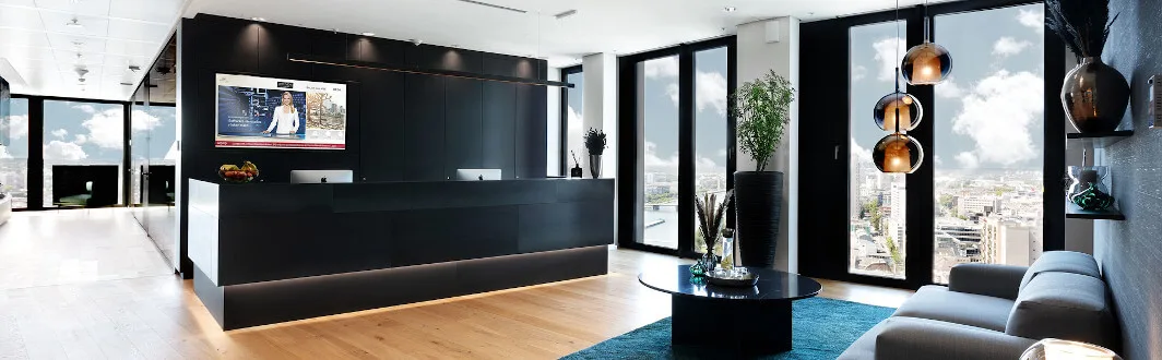 Moderne Empfangshalle mit dunklem Tresen, großem Bildschirm, Sofa, Teppich, eleganten Leuchten und bodentiefen Fenstern mit Stadtblick.