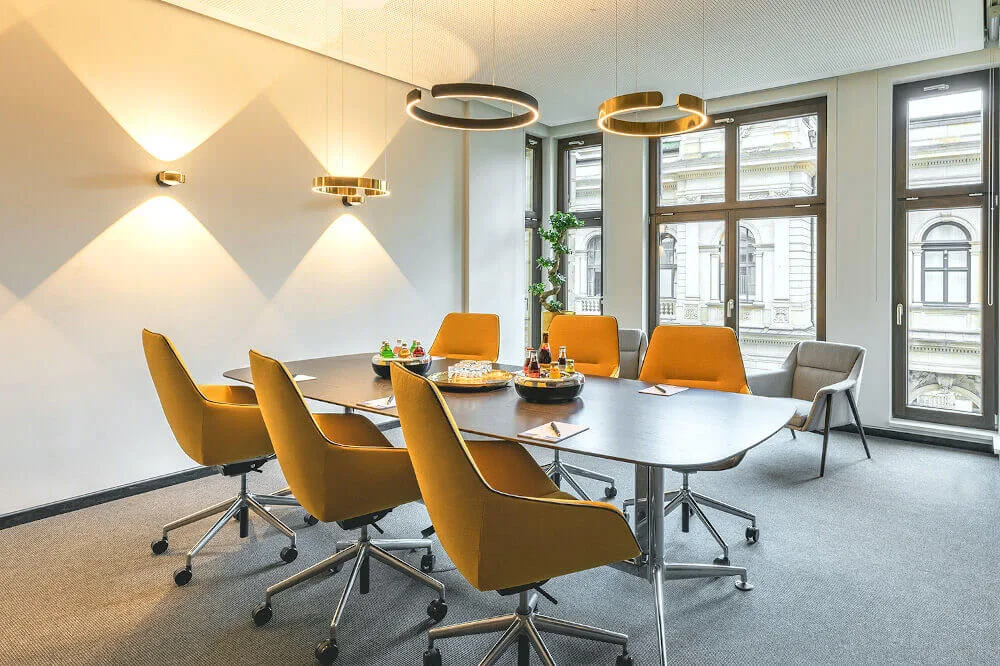 Moderner Konferenzraum mit großem Besprechungstisch, ergonomischen gelben Stühlen, stilvollen Pendel- und Wandleuchten sowie hohen Fenstern mit Blick auf eine Stadtansicht.