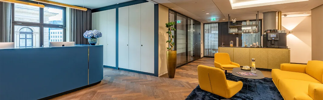Moderner Büroempfang mit blauem Rezeptionstresen, Blumen, gelben Lounge-Sitzmöbeln und Kaffeebar. Parkettboden und Glaswände.