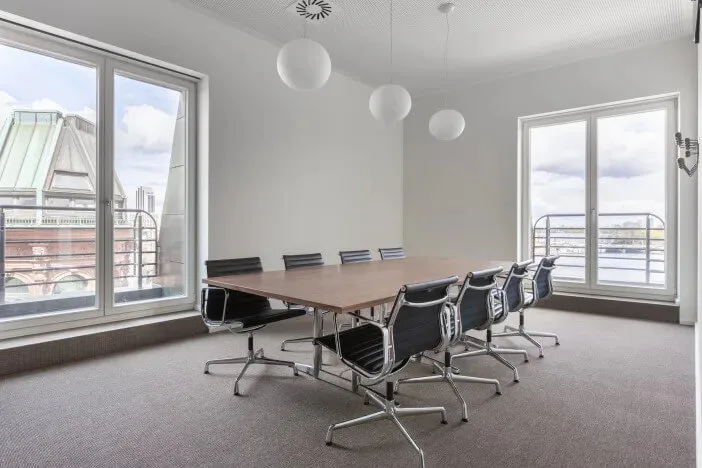 Moderner Konferenzraum mit langem Holztisch, schwarzen Stühlen, grauem Teppichboden und großen Fenstern mit Stadtblick.