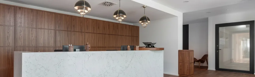 Moderne Rezeption mit weißer Marmortheke, hinter der eine Holzpaneelwand und drei stilvolle Pendelleuchten hängen. Rechts eine Glastür zu einem Flur.