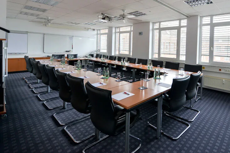 Großer Konferenzraum mit U-förmigem Tisch, vielen schwarzen Stühlen, Whiteboards, großen Fenstern und Teppichboden.