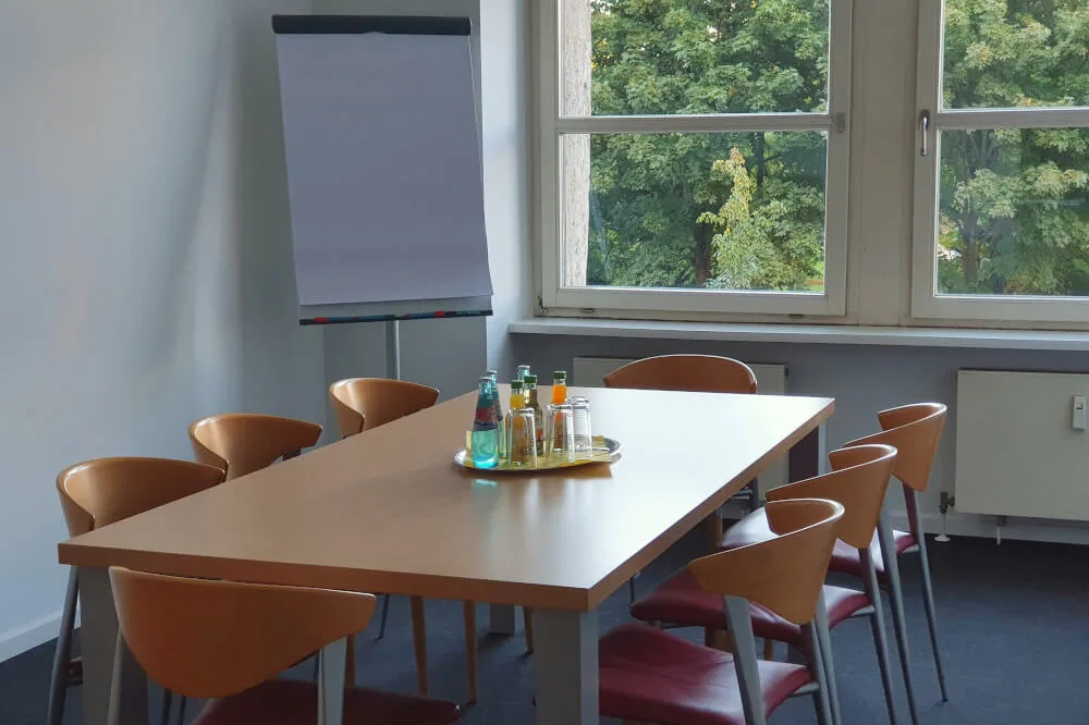 Besprechungsraum mit großem Holztisch, Stühlen, Flipchart, Getränken auf Tablett und Fenster mit Blick auf Bäume.