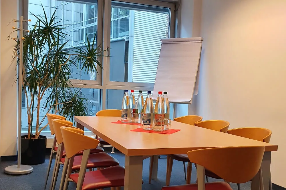 Moderner Konferenzraum mit Holztisch, orangefarbenen Stühlen, Wasserflaschen, Flipchart und großer Pflanze vor einem Fenster.