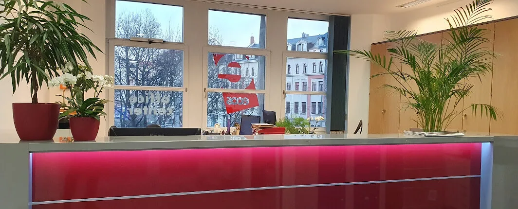 Rezeptionstresen mit leuchtend pinker Front, Topfpflanzen und großen Fenstern, die eine Stadtansicht zeigen.