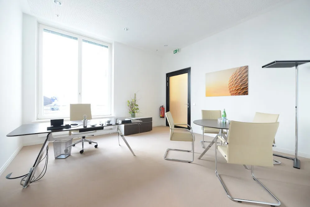 Helles, modernes Bürozimmer mit Schreibtisch, Bürostuhl, Besprechungstisch und vier Stühlen, großem Fenster und weißer Einrichtung.