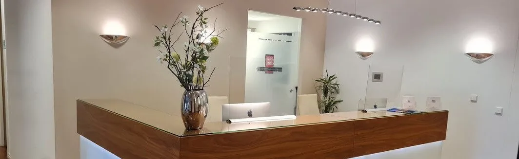 Moderne Rezeption mit Holztresen, Glasplatte, Vase mit Blumen, iMac und Wandleuchten in hellem Raum