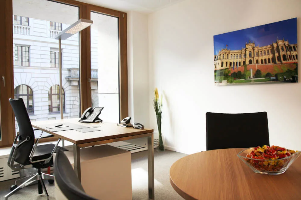 Modernes Büro mit Holzschreibtisch, Telefon und Stühlen, großem Fensterblick auf ein historisches Gebäude und Bild an der Wand.