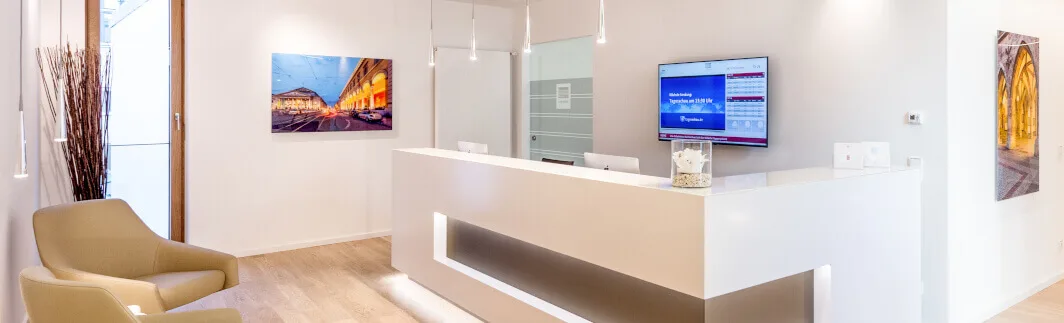 Moderner, heller Empfangsbereich mit weißer Rezeptionstheke, zwei beige Sesseln, Bildschirm, PCs und Kunst an den Wänden.
