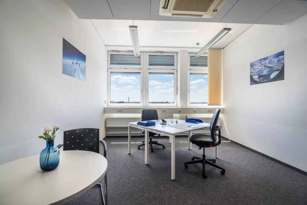 Helles, modernes Büro mit Besprechungstisch, Stühlen, grauem Teppich und großen Fenstern mit weitem Ausblick. Flugzeugbilder an der Wand.