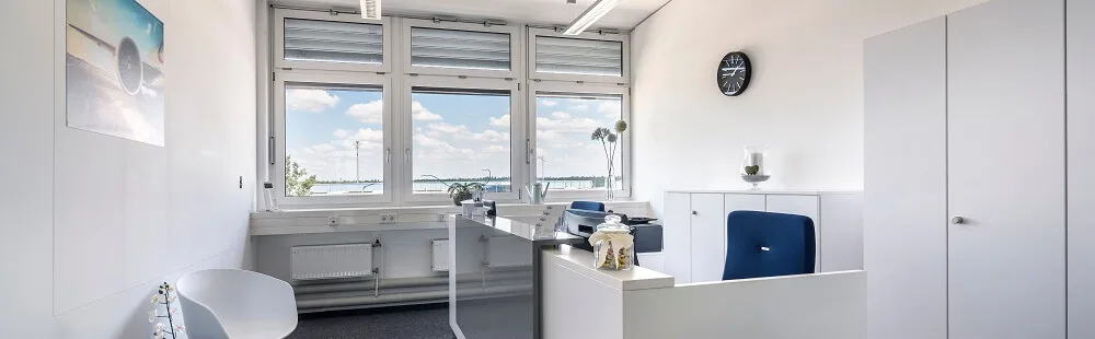 Moderne, helle Büroeinrichtung mit Empfangstheke, blauem Bürostuhl, weißen Schränken und großen Fenstern mit Seeblick.