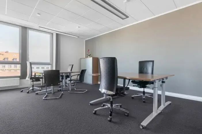 Mehrere höhenverstellbare Bürotische und ergonomische Drehstühle in einem modernen Büro mit grauen Wänden, Teppichboden und großen Fenstern.