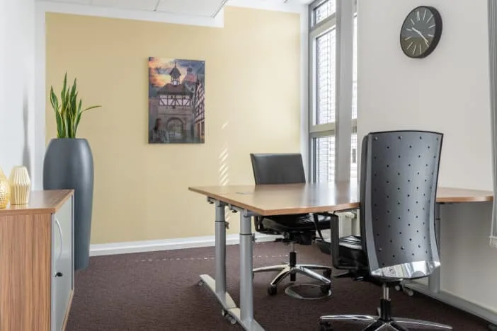 Heller Büroraum mit Holzschreibtisch, zwei Bürostühlen, einem Pflanzgefäß mit Grünpflanze und einem Bild an der hellgelben Wand. Ein Fenster und eine Wanduhr sind ebenfalls sichtbar.
