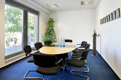 Konferenzraum mit ovalem Tisch, schwarzen Stühlen, blauem Teppich, großen Fenstern zum Balkon und gerahmten Bildern.