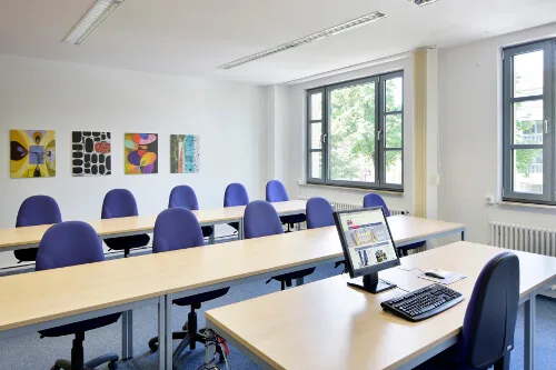 Moderner Seminarraum mit hellen Tischen, violetten Stühlen, vier bunten Bildern an der Wand und zwei großen Fenstern. Ein Computer steht auf einem Tisch.