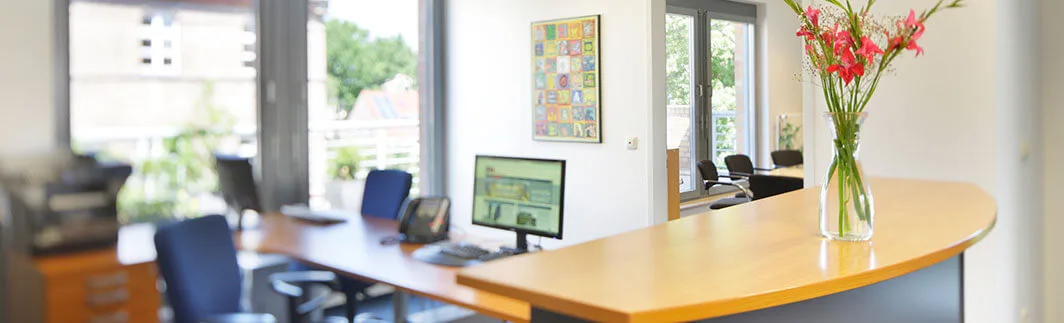 Heller moderner Bürobereich mit Empfangstheke, roten Blumen, Schreibtischen, Computern und blauen Stühlen. Große Fenster im Hintergrund.