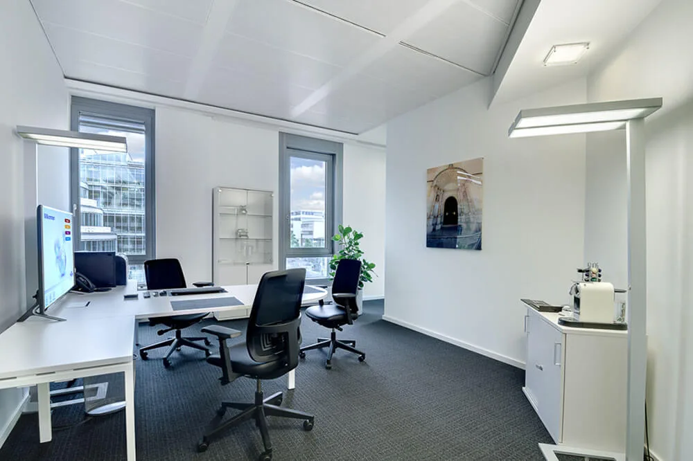 Modernes, helles Büro mit L-Schreibtisch, zwei Bürostühlen, grossen Fenstern, Zimmerpflanze, Wandbild und Kaffeemaschine.