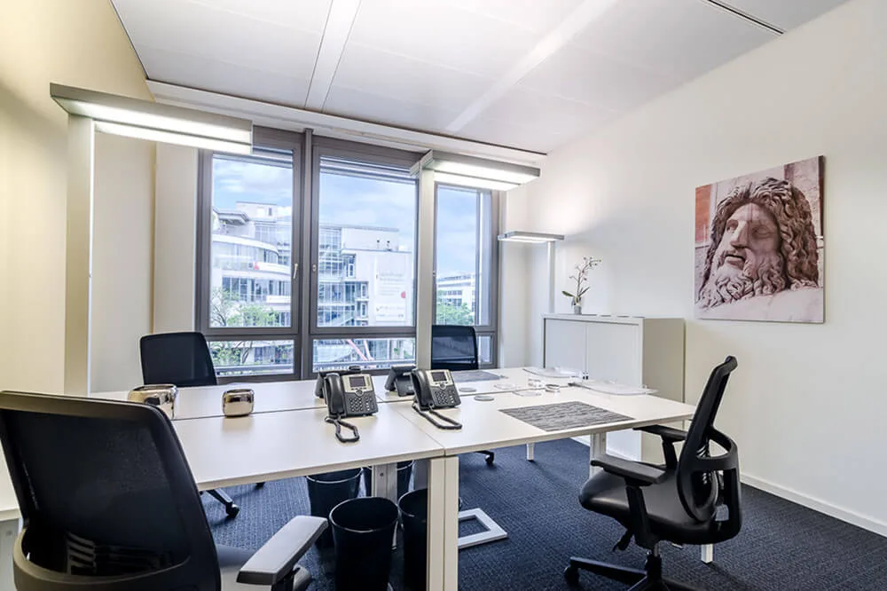 Modernes Büro mit zwei Arbeitsplätzen, schwarzen Bürostühlen, Telefonen, Fensterblick auf Gebäude und einem antiken Büstenbild.