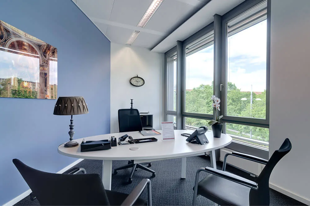 Modernes Bürozimmer mit ovalem Besprechungstisch, zwei Stühlen, Fensterfront mit Blick ins Grüne und Wanddekoration.
