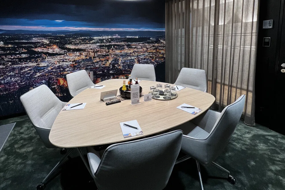 Moderner Besprechungsraum mit asymmetrischem Holztisch und sechs Stühlen. Wandtapete zeigt nächtliche Stadtansicht.