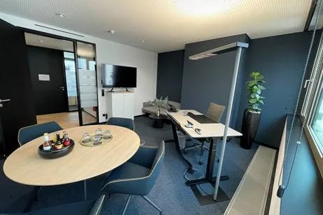 Moderner Besprechungsraum mit ovalem Holztisch, dunkelblauen Stühlen, Schreibtisch, Fernseher und Fenster mit Jalousie