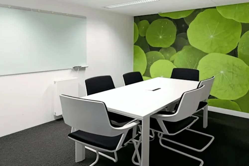 Moderner Besprechungsraum mit weißem Tisch, schwarzen Stühlen, Whiteboard und einer grünen Blatt-Fototapete an der Wand.