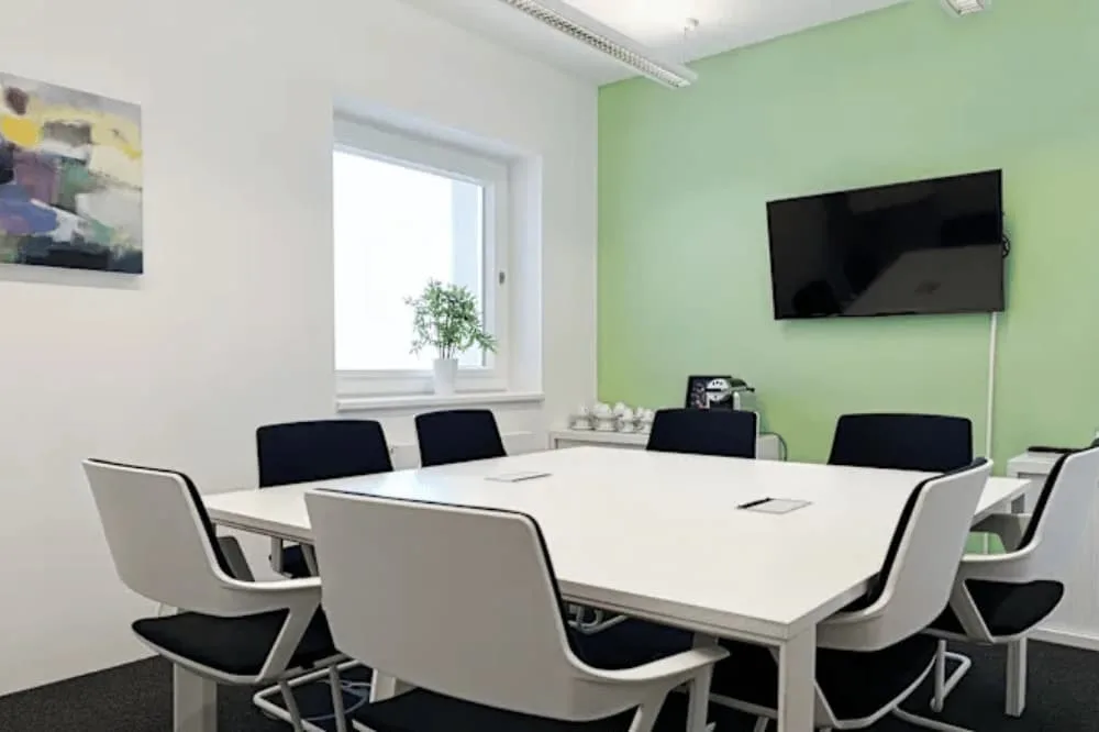 Moderner Besprechungsraum mit weißem Tisch, mehreren Stühlen, grün-weißen Wänden, Fenster, Pflanze, TV und Gemälde.