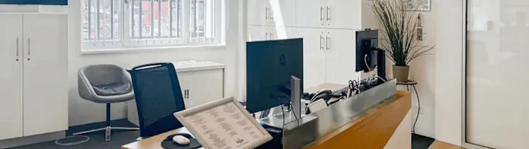 Moderner Empfangsbereich mit einer Holztheke, zwei Computermonitoren, Bürostühlen, weißen Schränken und einem Fenster mit Jalousien