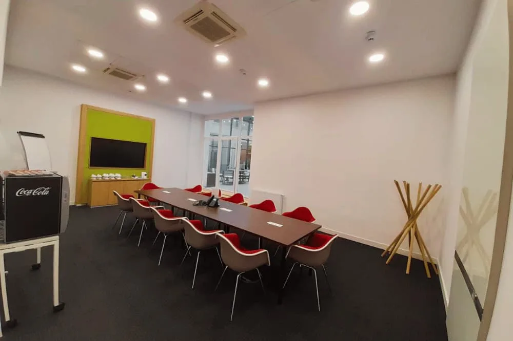 Moderner Konferenzraum mit langem Tisch, roten Stühlen, großem Bildschirm, Coca-Cola Kühlschrank und Flipchart.