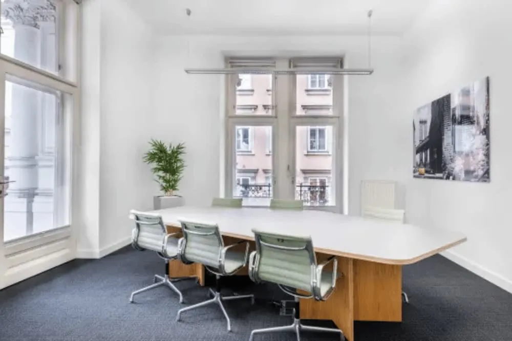 Helles, modernes Besprechungszimmer mit großem Konferenztisch, sechs Stühlen, einer Pflanze und Fenstern zu einer Stadtansicht.